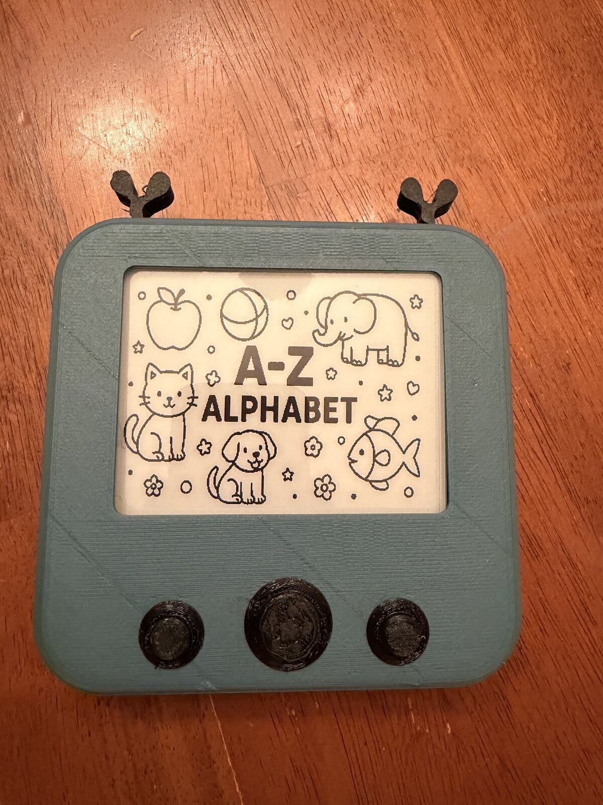 A green prototype with small antlers, three black front buttons, and an alphabet book on its display
