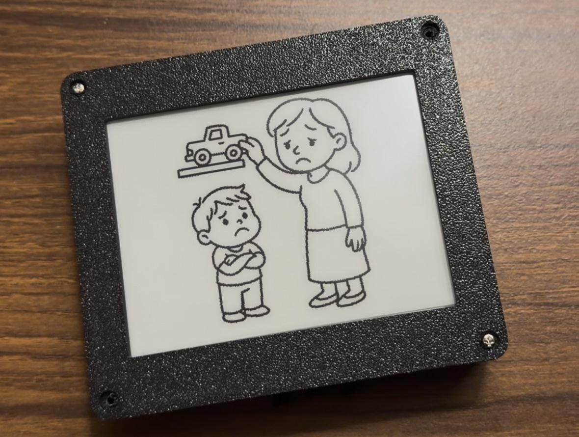 A Tinku in a black 3D-printed case with screwed corners, showing a line drawing of a mother holding a toy truck above a child