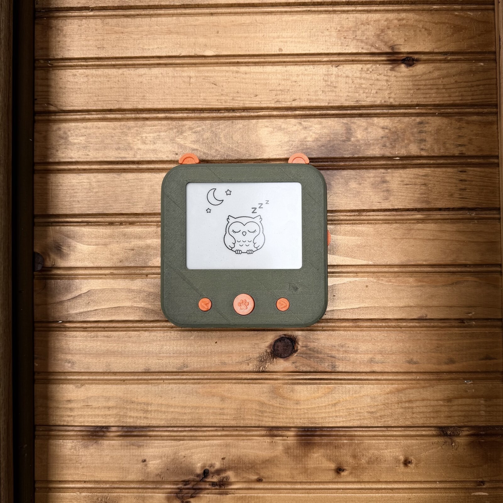 The finished Tinku: a rounded green case with orange buttons and bear ears, showing a sleeping owl and a crescent moon on its display